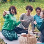 Pretty young women are taking selfie with smartphone during picnic in park sitting on grass, posing and laughing having fun. Nature, friendship and modern technology concept.