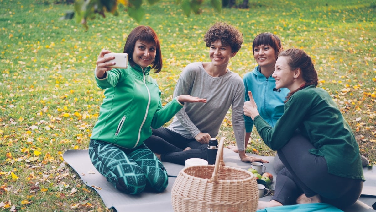 Pretty young women are taking selfie with smartphone during picnic in park sitting on grass, posing and laughing having fun. Nature, friendship and modern technology concept.