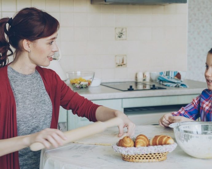 Young mother rolling dough and talking to her little cute daughter while cooking in the kitchen on weekend. Family, food, home and people concept