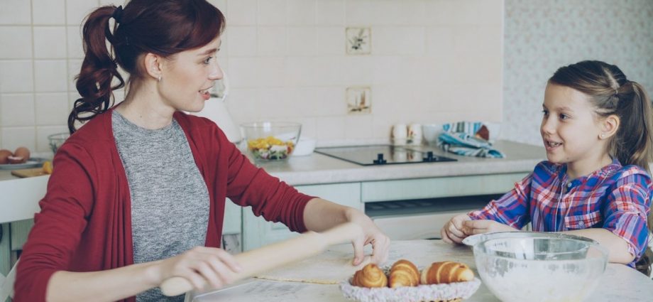 Young mother rolling dough and talking to her little cute daughter while cooking in the kitchen on weekend. Family, food, home and people concept
